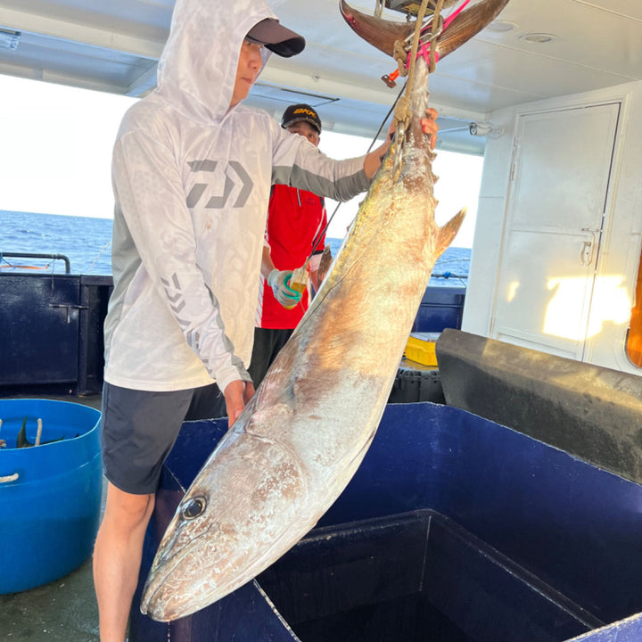 A person on a fishing boat holds a large fish hoisted by a hook, wearing a light-colored long-sleeve shirt and a cap. In the background, another person in a red shirt is visible, along with fishing equipment and containers. Sunlight streams through the boat's windows, and the ocean is visible outside.